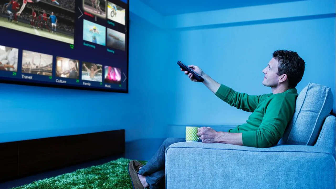 Man on a sofa pointing a remote at a large TV showing a grid of programme categories