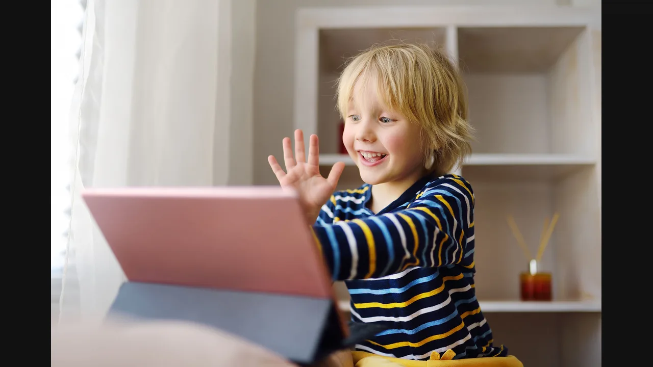 Smiling child waving at a tablet propped up on a table at home